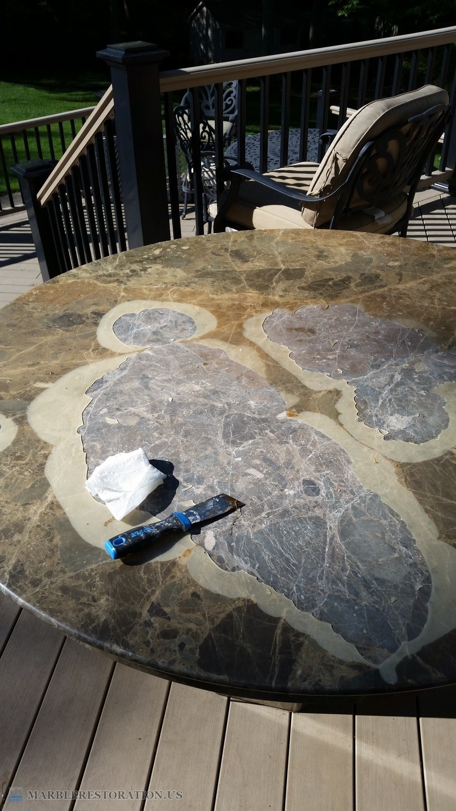 Peeling resin coating on a round Emperador marble table, before Round brown marble table on a deck, the old resin coating peeling and scraped off in large patches, a putty knife on top