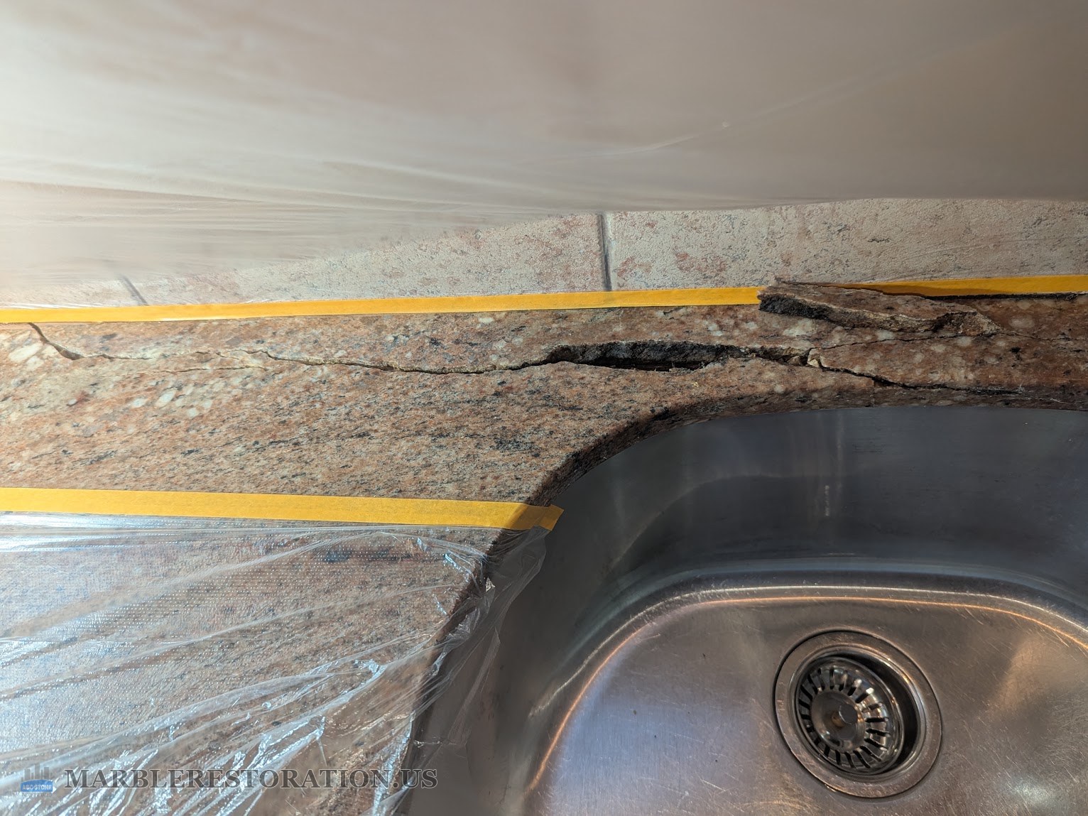 Pink granite kitchen countertop with a long open crack between an undermount stainless sink and the tile backsplash, plastic and yellow tape around it