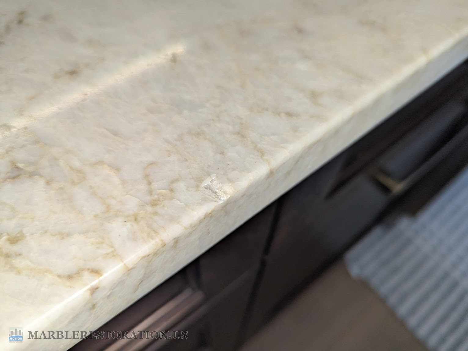 Chip on the edge of a leathered quartzite kitchen countertop, before Leathered white quartzite kitchen countertop with tan veining, a small chip flaked off the top edge, dark wood cabinets below
