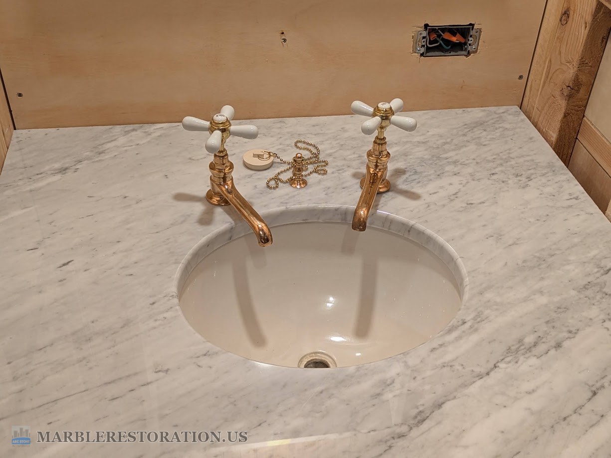 Two brass cross-handle faucets set in square mortises on a Carrara marble vanity top, the drain plug stanchion between them