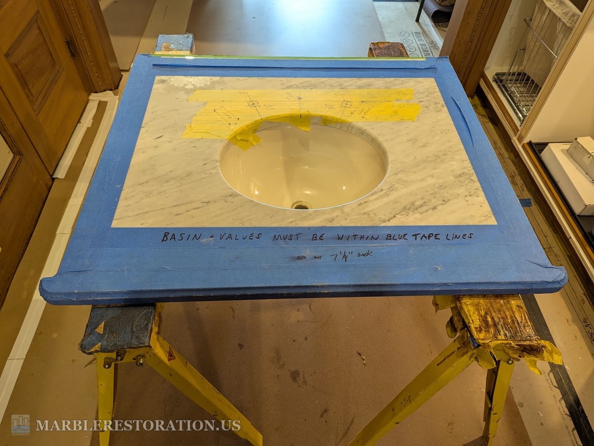 Carrara marble vanity top with an undermount sink basin on sawhorses, faucet hole layout marked on yellow tape