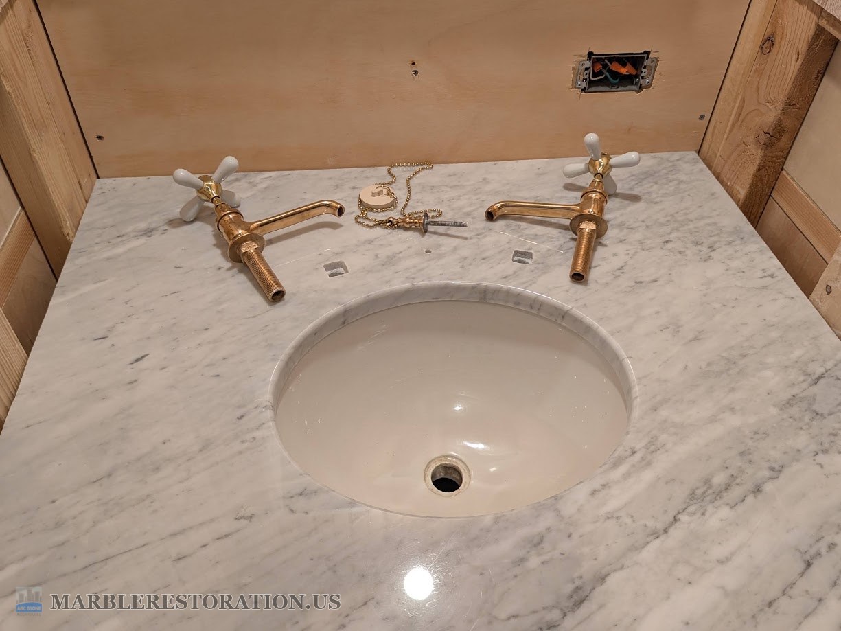 Carrara marble vanity top with two square faucet mortises and a drain plug stanchion hole, brass cross-handle faucets beside them