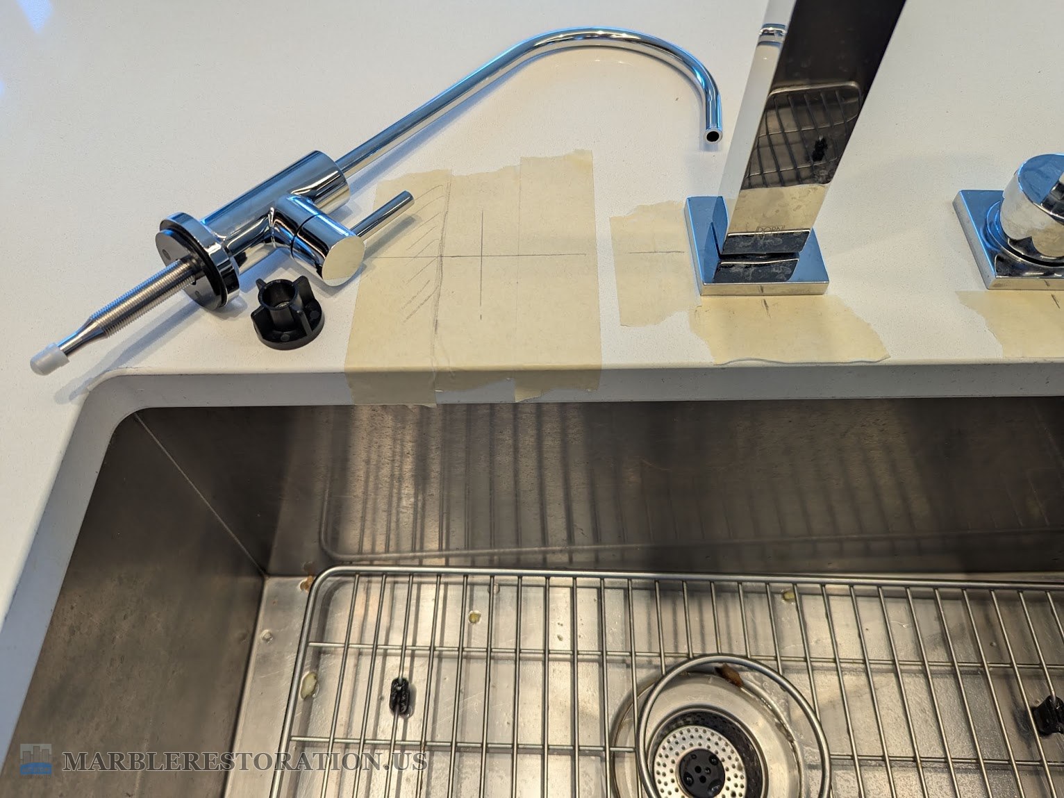 Quartz countertop marked for a water filter faucet hole, before Masking tape with a pencil cross on a white quartz countertop, a chrome filter faucet and its black mounting nut lying beside it