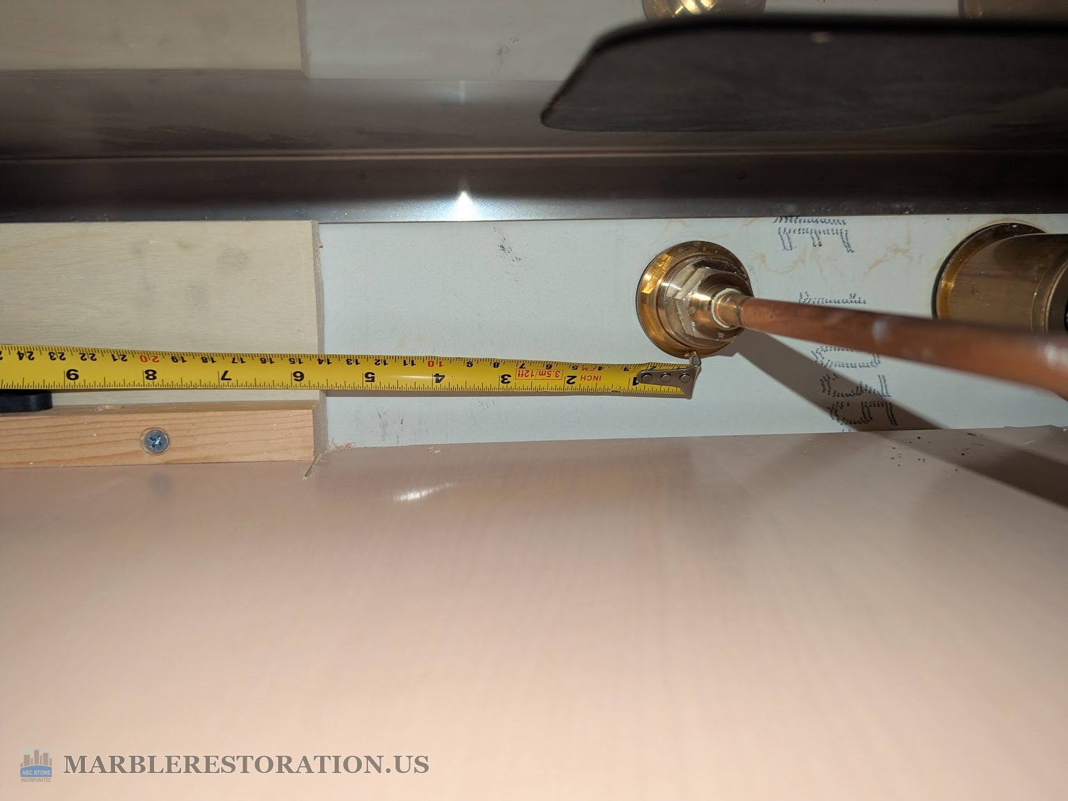 Measuring under the quartz countertop past the wooden support A tape measure under a quartz countertop running from a brass faucet supply fitting to the drilling spot, with the unexpected wooden support at the left